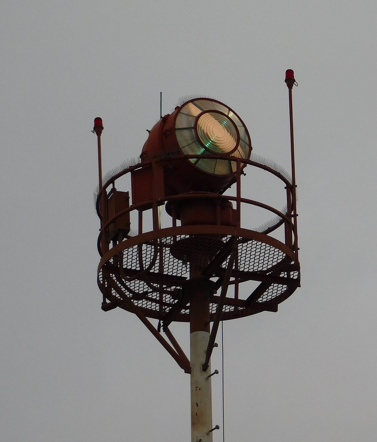 A large round beacon lamp with a glass front on a railed platform at the top of a mast, against a grey sky.