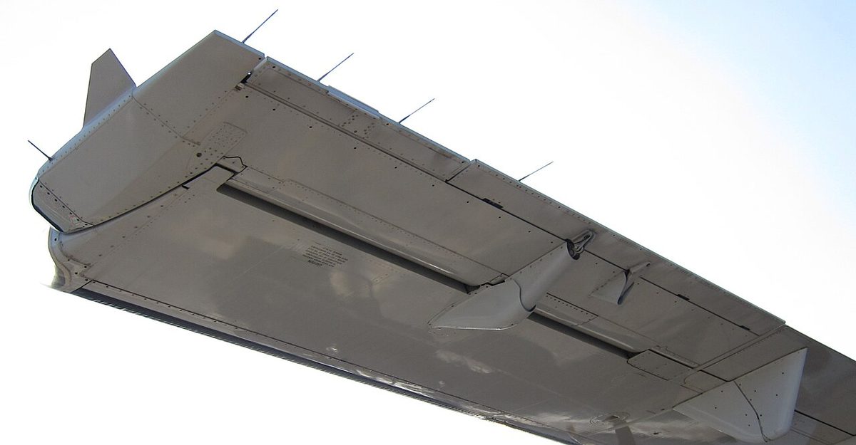 The trailing edge of a turboprop's left wing seen from behind and below, showing the aileron with two narrow tabs along its rear edge.