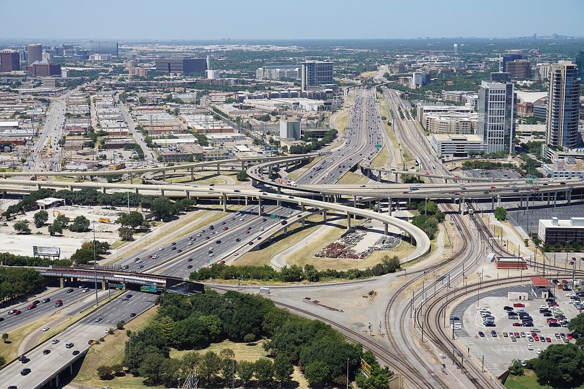 A high view over a large motorway interchange in Dallas, with curving flyover ramps linking multi-lane motorways and railway lines running alongside.