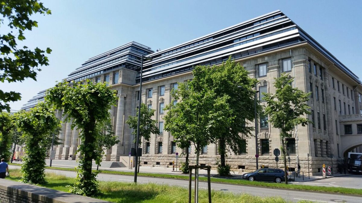 A large stone-faced office building in Cologne with modern glazed storeys added on top, seen behind a row of trees.