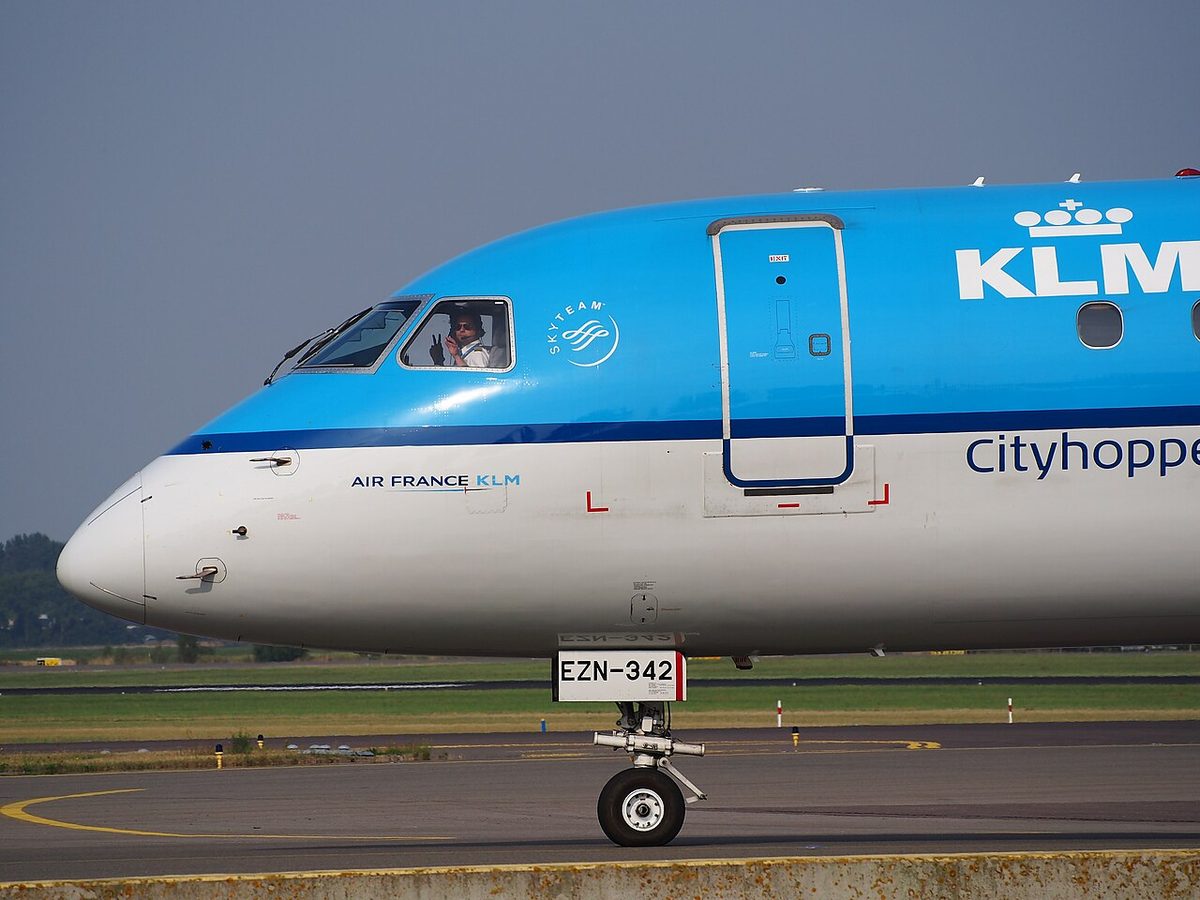 The nose of a blue and white KLM Cityhopper Embraer 190 taxiing, a pilot looking out of the side window, with L-shaped probes below the cockpit windows and a wiper on the windshield.