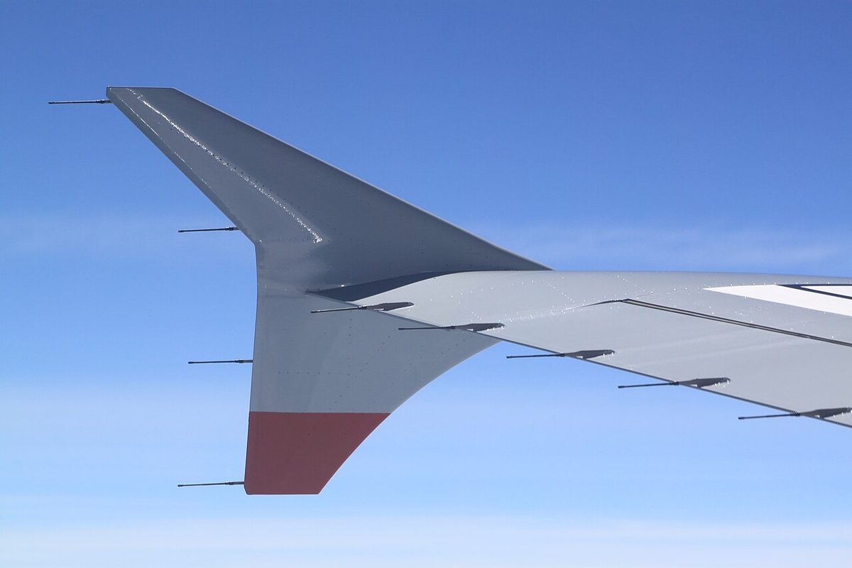 Wing tip of an airliner in flight with a small arrowhead-shaped plate rising above and hanging below the tip, and thin rods sticking out behind its edges and the wing's trailing edge.