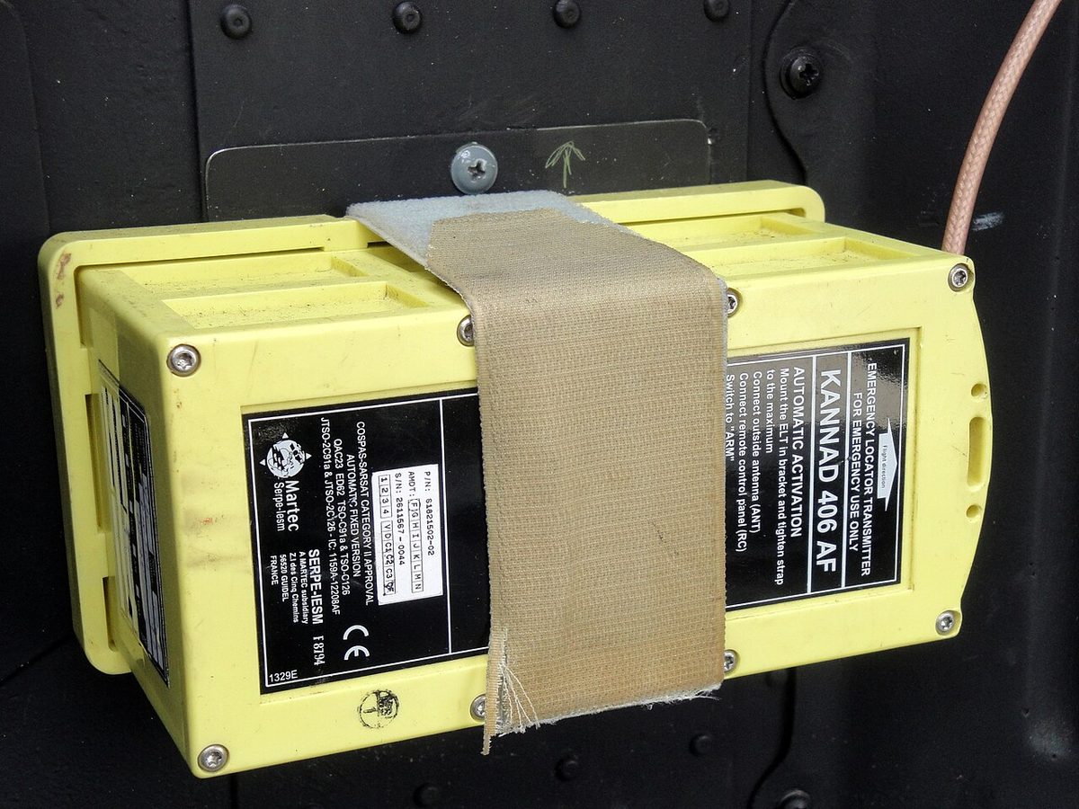 A yellow box-shaped emergency locator transmitter held to its mounting tray by a fabric strap, with an antenna cable leading away.