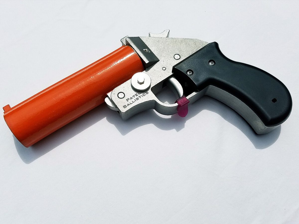 A 26.5 mm flare pistol with an orange barrel, a bare metal frame and a black grip, lying on a white surface.