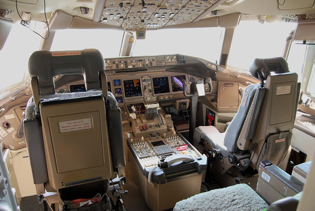 The flight deck of a Boeing 777 seen from behind the pilots' seats, with large display screens across the instrument panel and a centre pedestal between the seats.