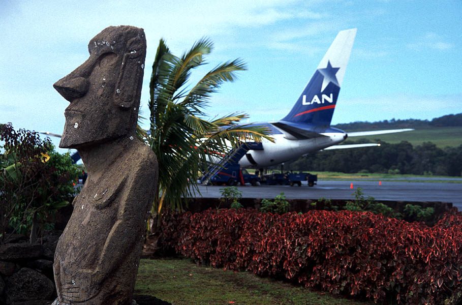 A moai statue beside the apron at Mataveri airport on Easter Island, with a LAN airliner parked behind a hedge and palm.