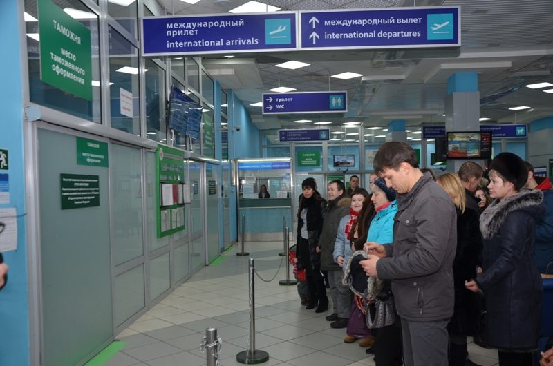 Travellers waiting in a terminal hall beside glazed customs booths, under signs in Russian and English for international arrivals and departures.