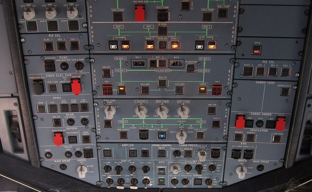 A cockpit ceiling panel of grey switch panels with rows of square pushbuttons, a few lit amber, red guarded switches and white rotary knobs.