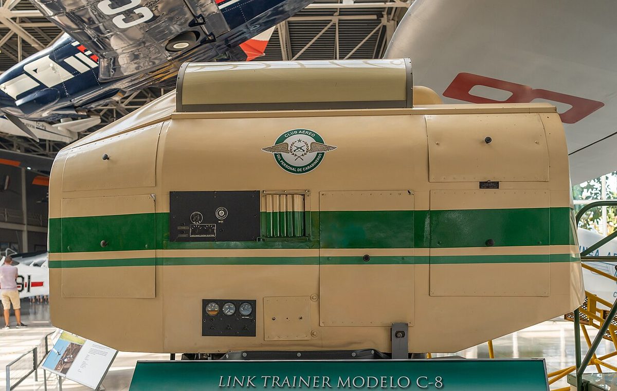 A Link Trainer, a small enclosed replica cockpit with stubby wings and a tail, mounted on a pedestal base.