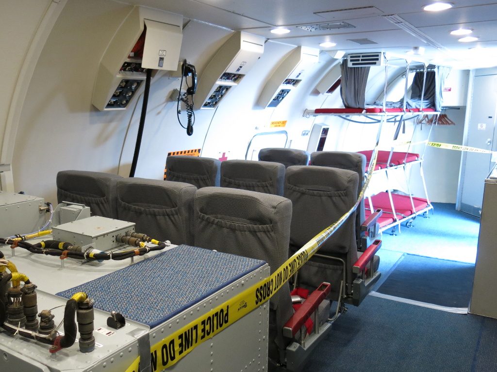 Rows of grey seats and three tiers of red bunks in an aircraft cabin, behind a yellow tape.