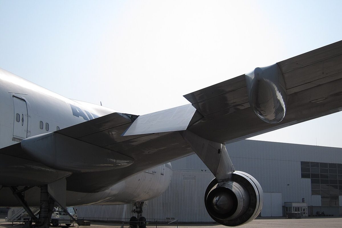 The underside of an airliner's wing seen from near the fuselage, with a panel deflected in the trailing edge between flap sections, inboard of an engine.