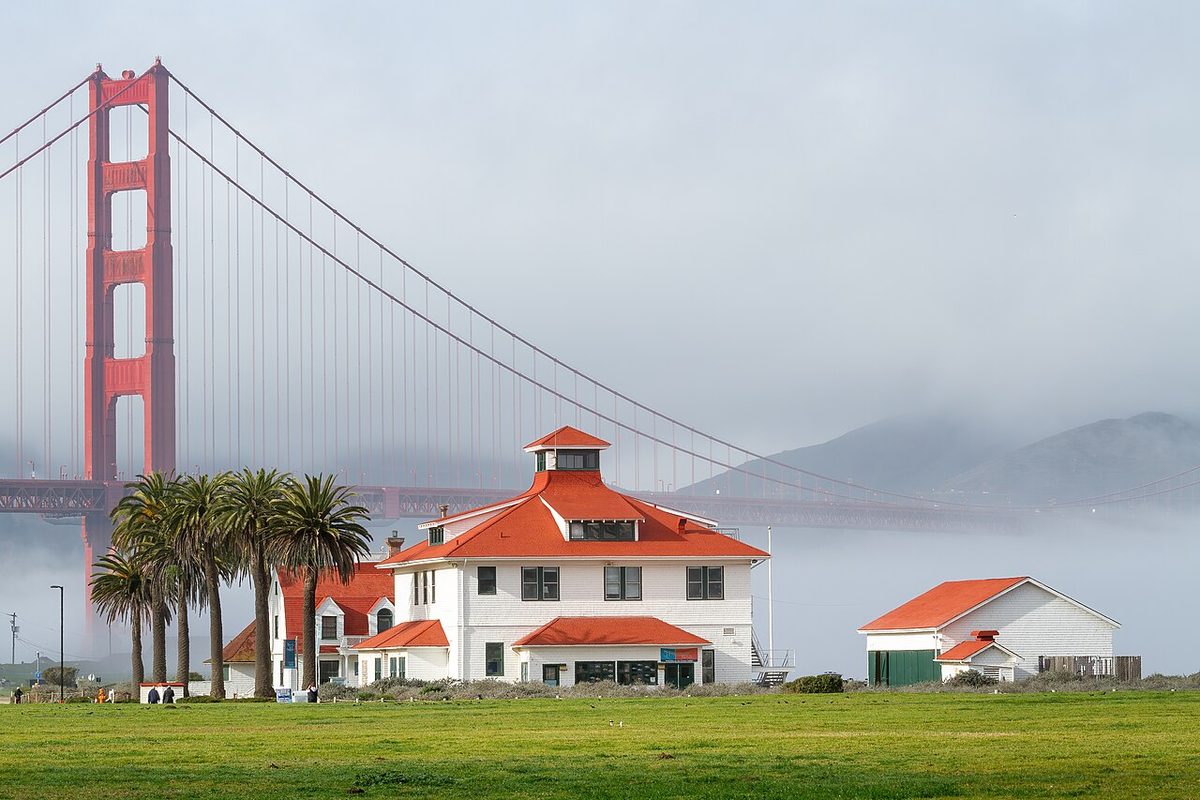 Fog lying over San Francisco Bay behind the old Crissy Field Coast Guard station, with a tower of the Golden Gate Bridge rising into it.