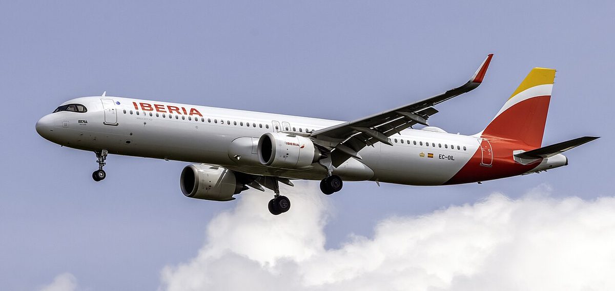 A long white airliner with a red and yellow tail on final approach, landing gear down, upturned wingtips and clouds below.