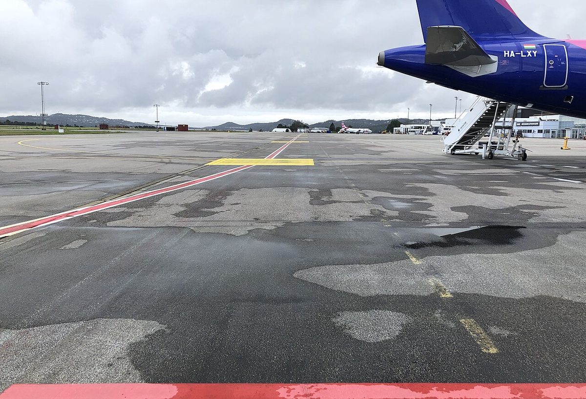 An airport apron seen from ground level, with a yellow painted block and red lines on patched tarmac, and an airliner's tail and boarding stairs on the right.