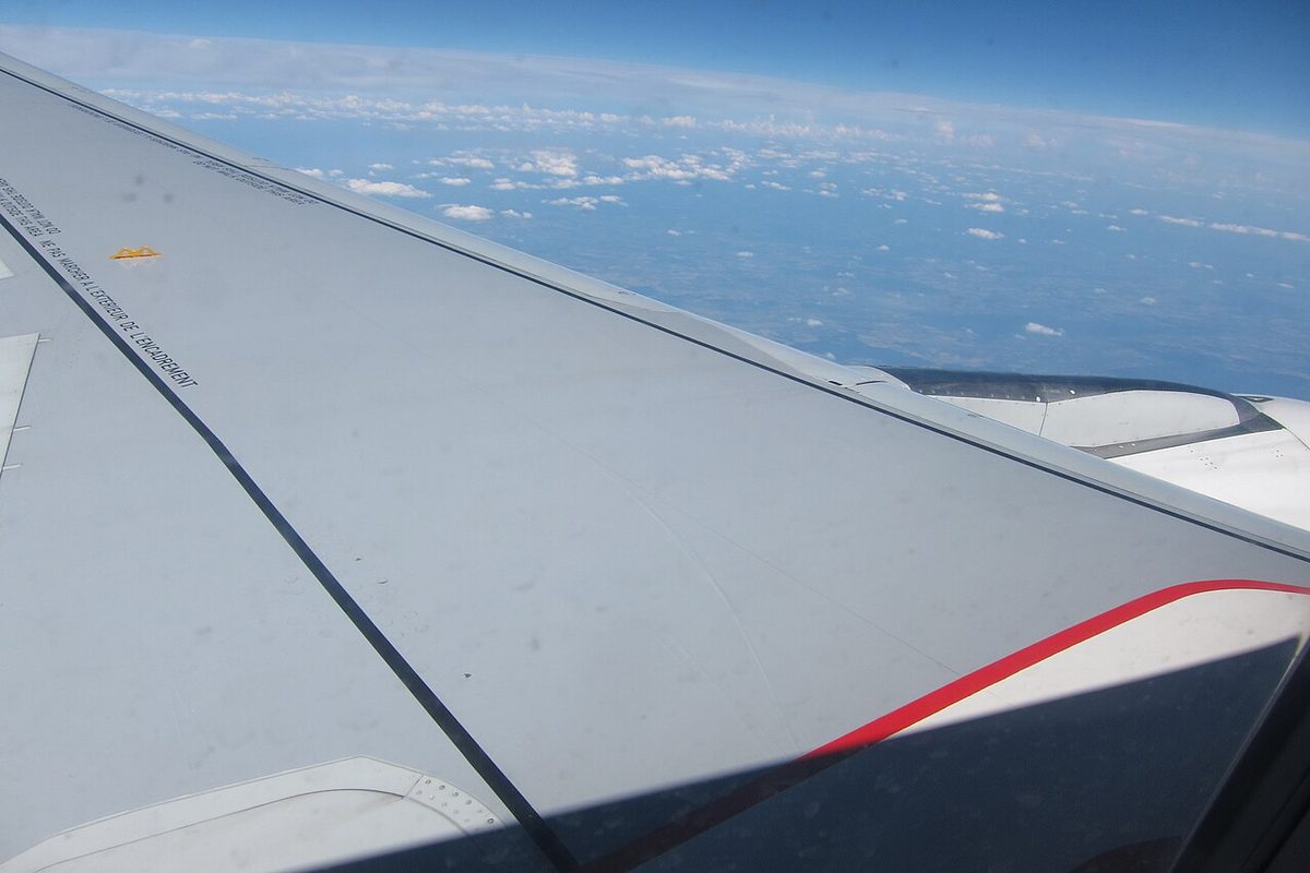View from a cabin window of an airliner wing in cruise, with a thin line of refracted light marking a shock wave standing on the upper surface.