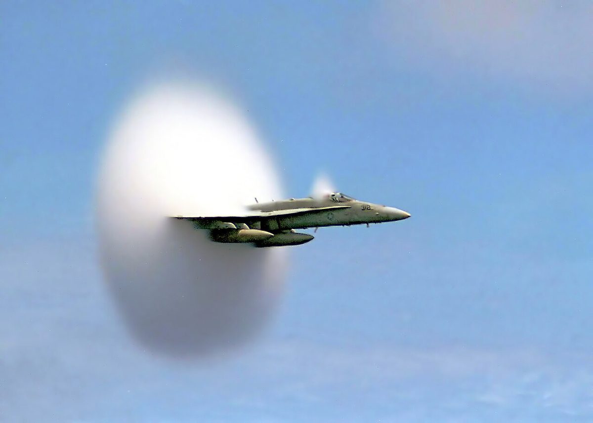 A US Navy F/A-18 Hornet flying low over the sea surrounded by a cone-shaped cloud of condensation.