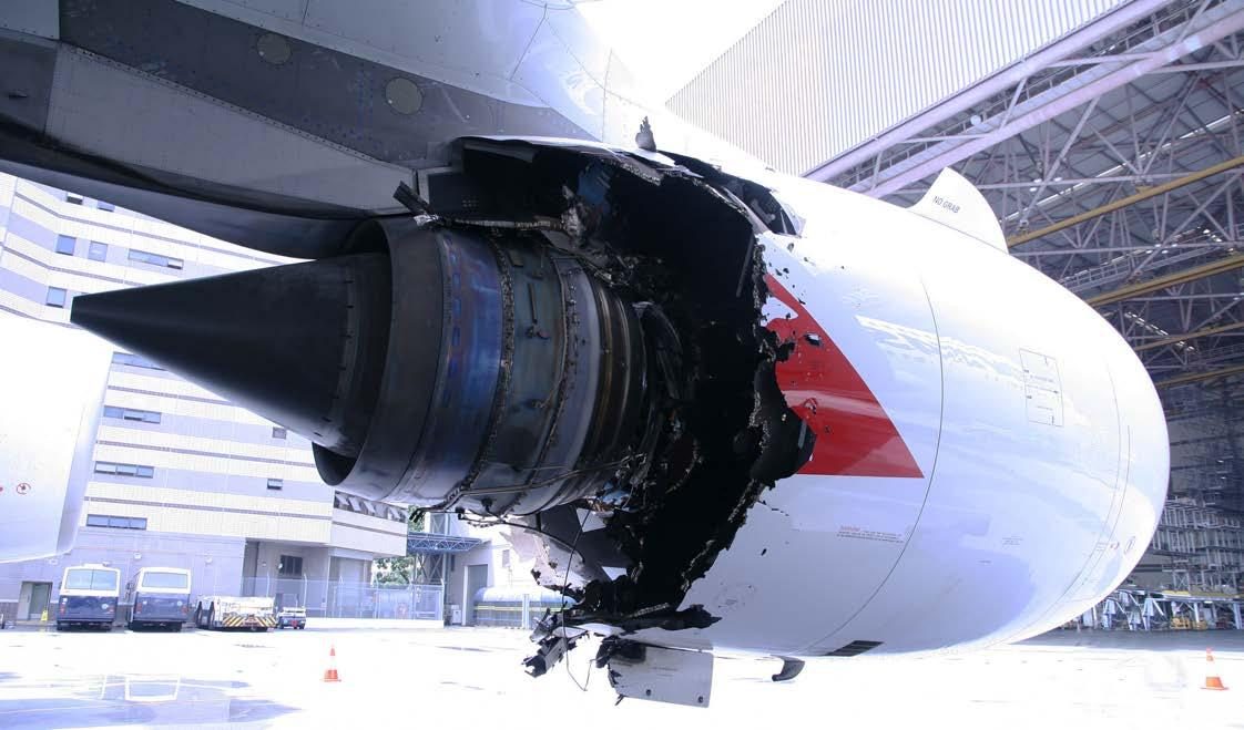 An airliner engine under a wing, seen from behind outside a hangar, with a large jagged hole torn through the rear of its nacelle.