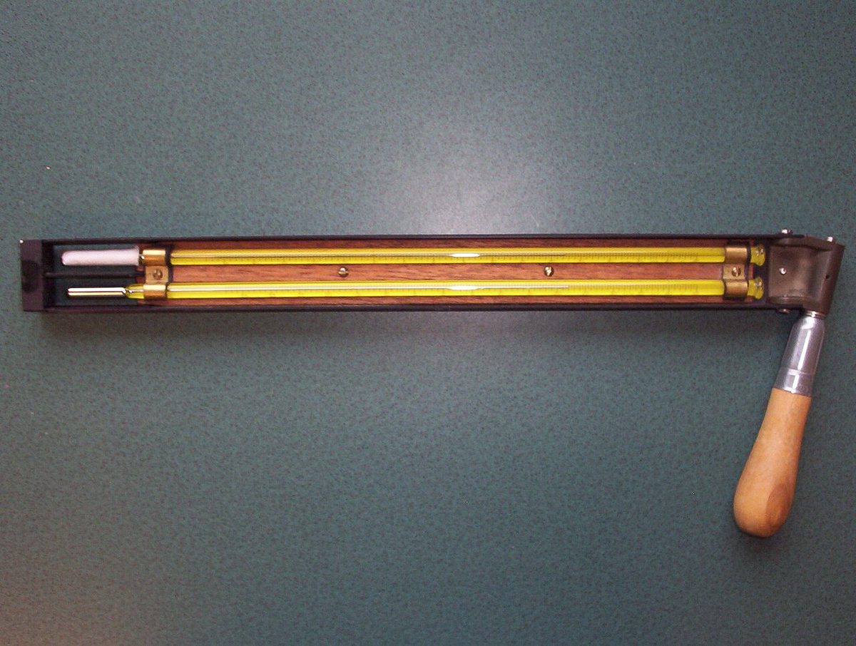 A sling psychrometer: two thermometers side by side in a frame that is whirled round by its wooden handle, one with a wetted sleeve over its bulb.