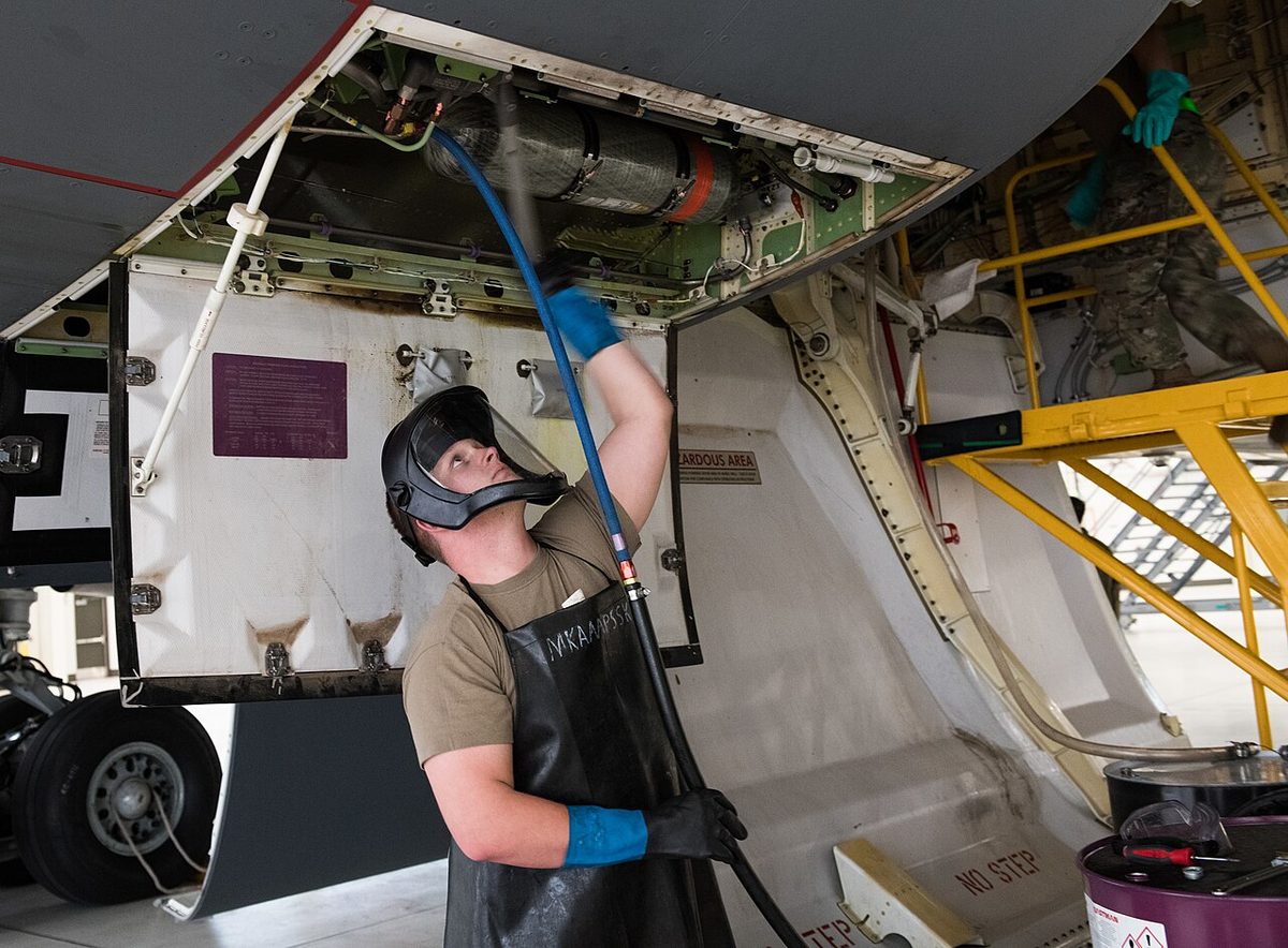 A maintainer in a face shield, apron and long gloves servicing the hydraulic system of a large aircraft through an open panel.