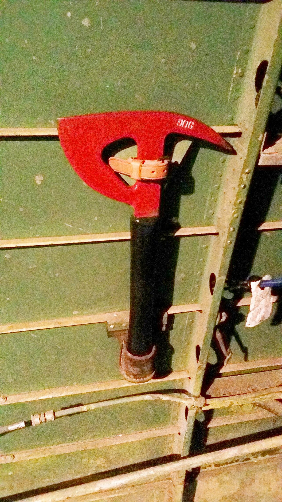 A red-headed axe with a dark handle clipped to the green-painted inside wall of an aircraft fuselage.
