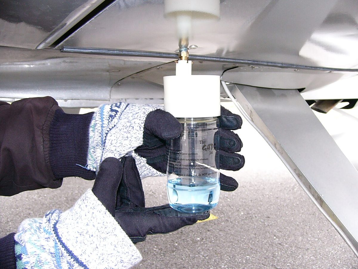 Gloved hands holding a clear jar pressed against a drain valve under an aircraft wing, the jar partly filled with blue fuel.