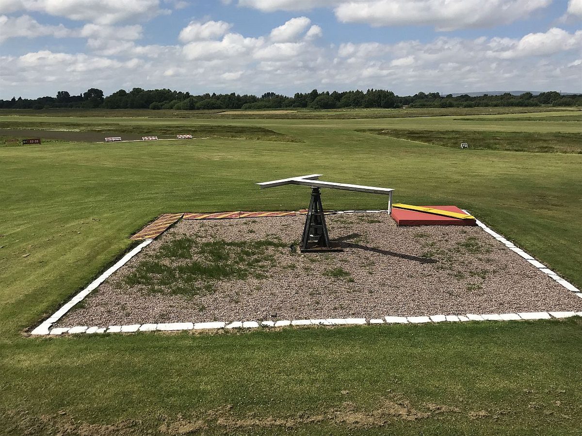 A gravel square edged in white on an airfield's grass, with a white T-shaped sign on a stand and red and yellow panels.