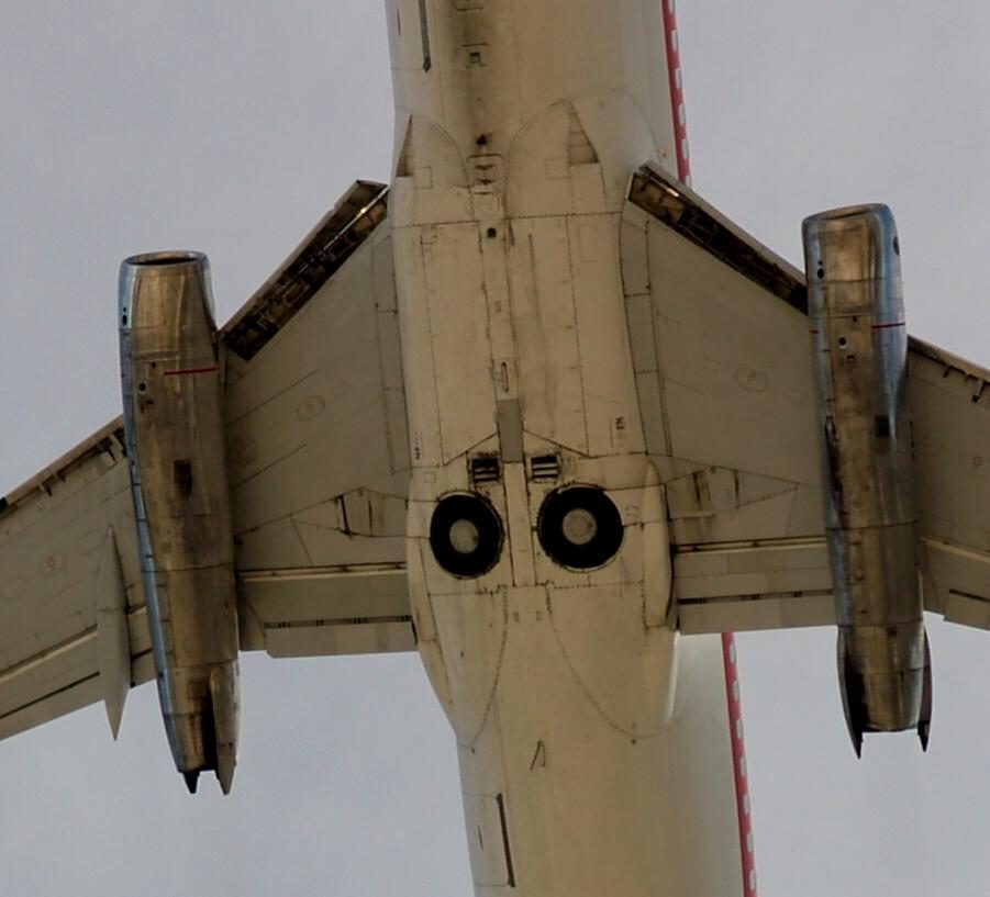 An airliner seen from directly below in flight with its gear up: two tyres sit uncovered in round openings in the belly between the wings.
