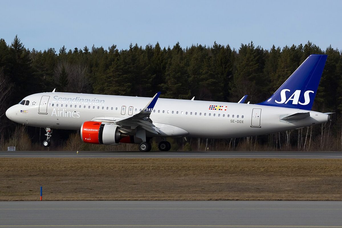 A white SAS Airbus A320neo with a dark blue tail and red engine cowls touching down on a runway, its nose wheel still in the air, with pine forest behind.