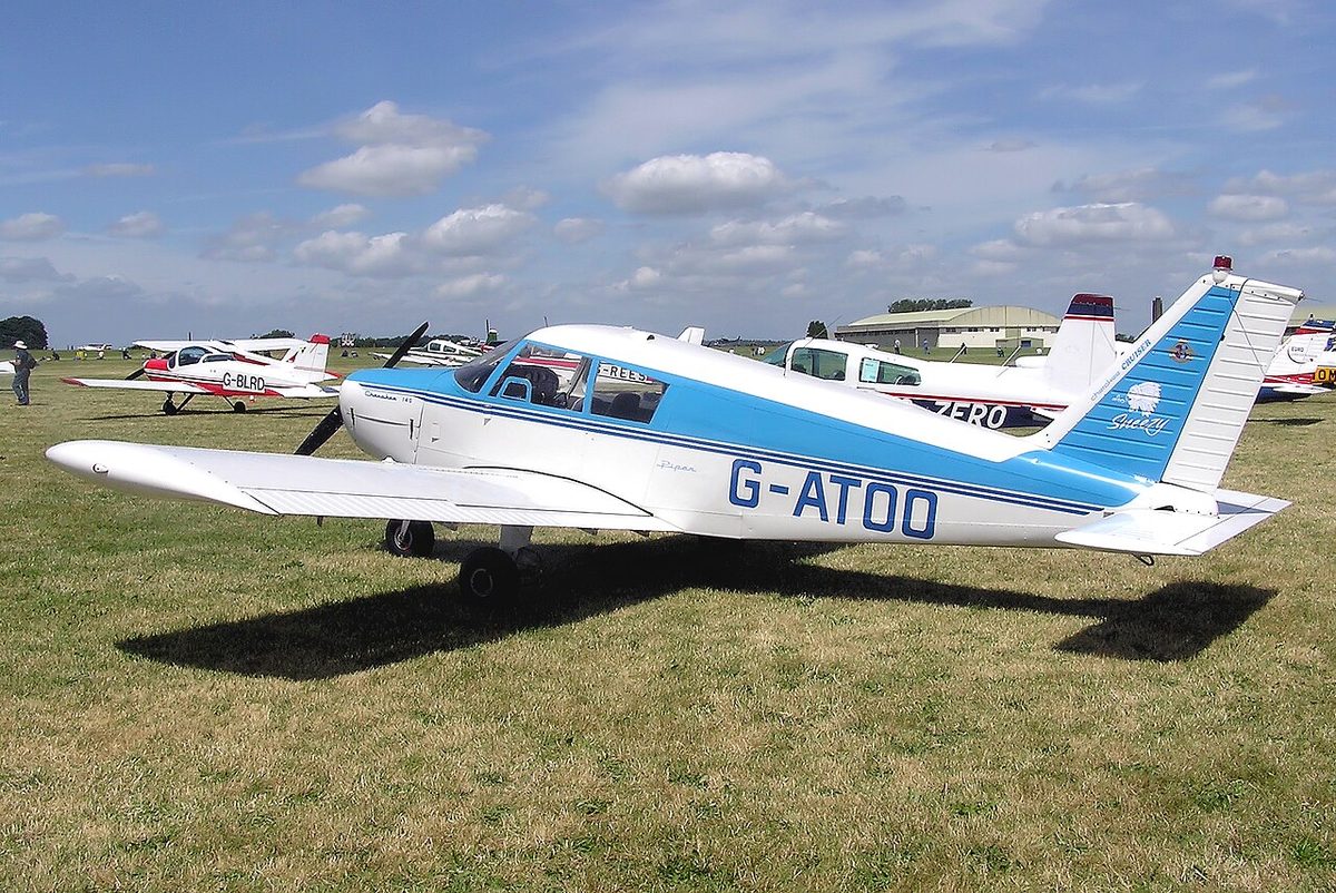 A white and blue low-wing Piper light aircraft registered G-ATOO parked on grass among other light aircraft.