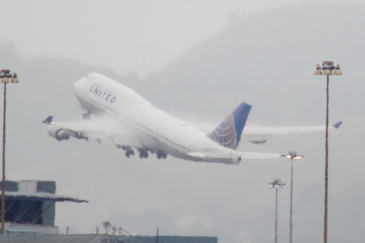 A Boeing 747 climbing away in rainy, humid air, with a white sheet of condensation over the upper surface of its wing.