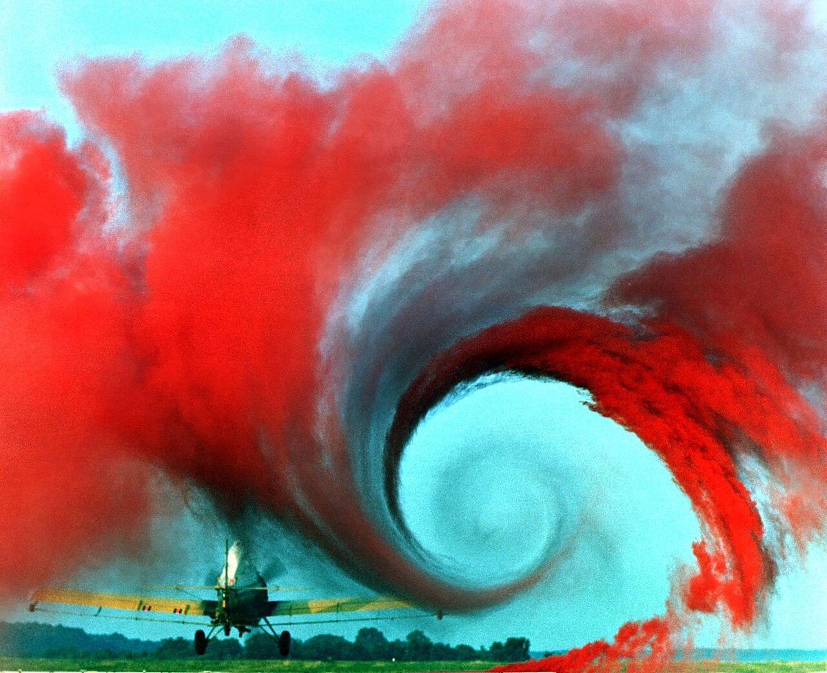 Coloured smoke rising from the ground is drawn into a large spiral behind the wingtip of a low-flying aircraft.