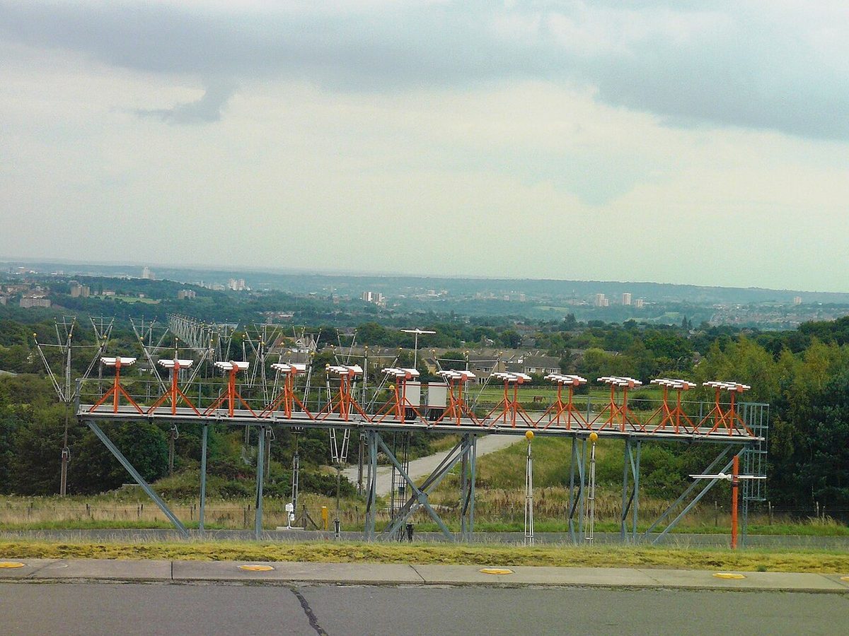 A localiser antenna array at Leeds Bradford Airport: a row of orange and white aerials on a raised steel platform beyond the runway, with houses and hills behind.