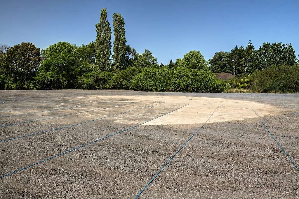 A large, weathered pad of rough paving with a paler concrete circle at its centre and blue lines radiating across it, trees behind.