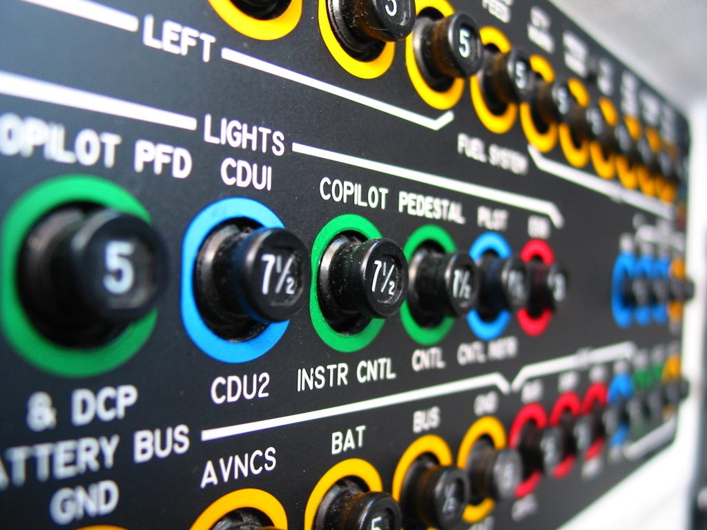 Close-up of rows of round push-pull circuit breakers with coloured collars and labels such as COPILOT PFD, CDU and BAT.