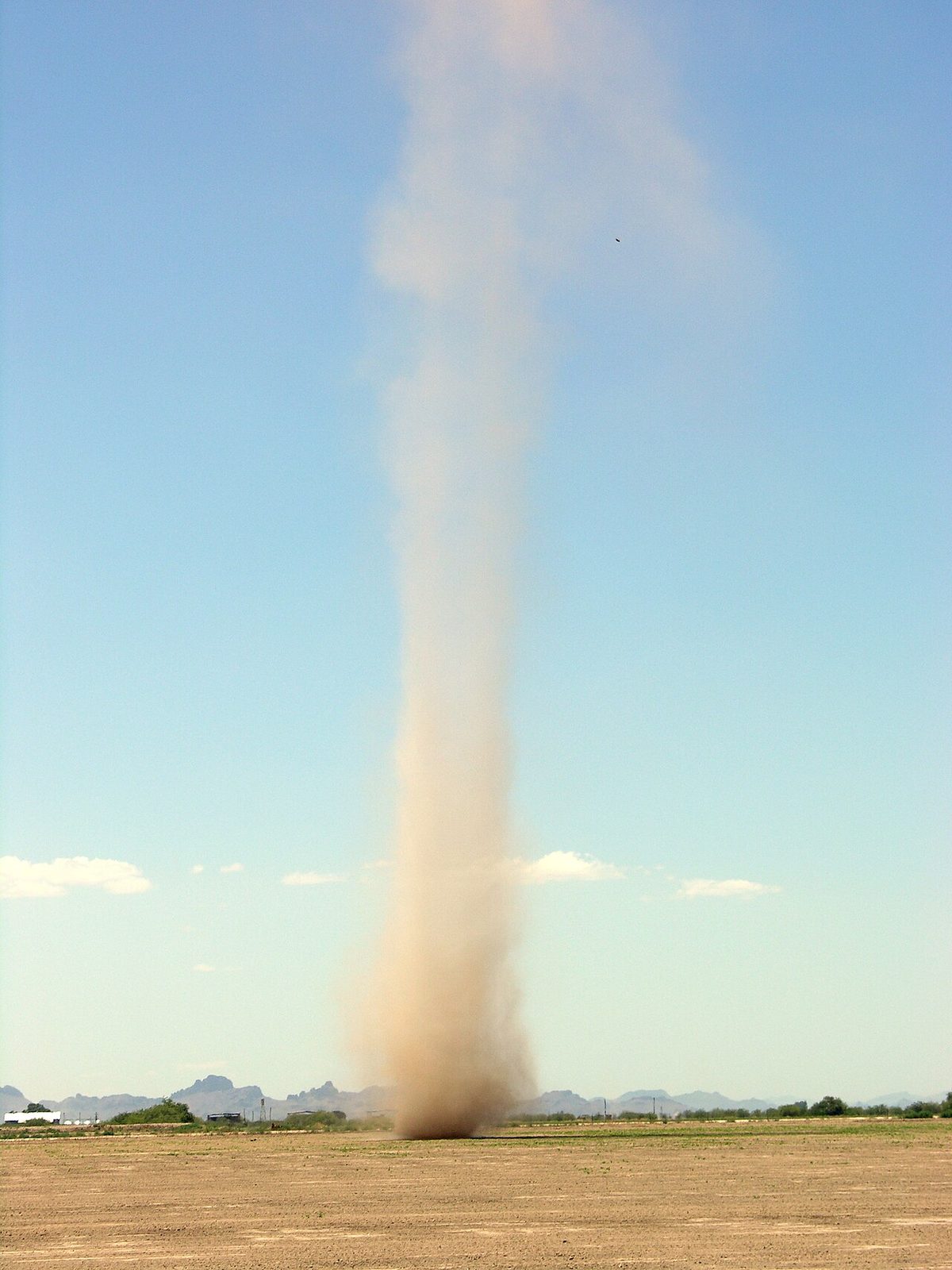 A tall column of dust spinning up from flat, dry ground under a clear sky.