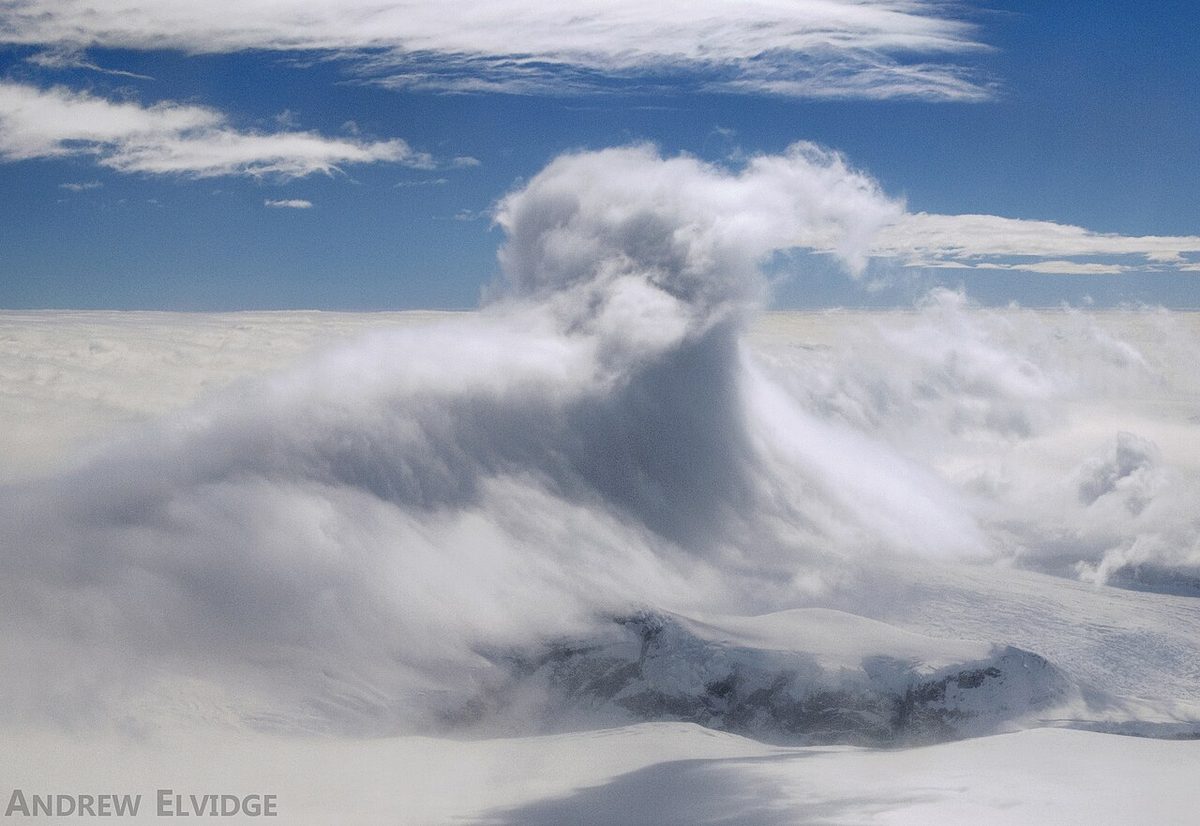 A billowing, overturning cloud above snow-covered slopes under a blue sky.