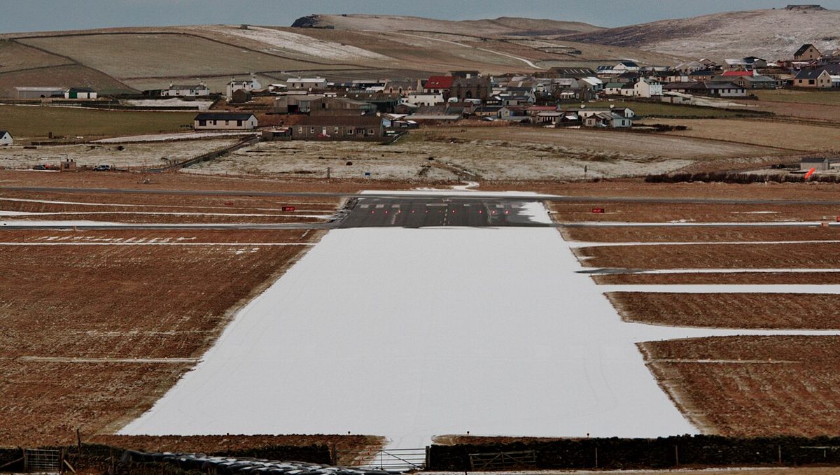 A small island airfield's runway covered in snow, with a village and snowy hills behind it.