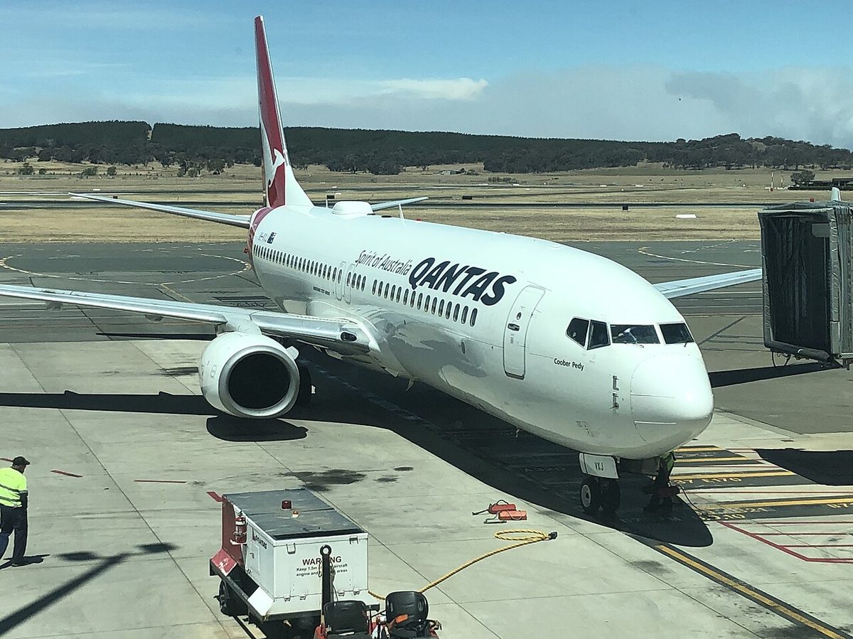 A white Qantas Boeing 737 with a red kangaroo tail parked at a jet bridge, with ground equipment around it.
