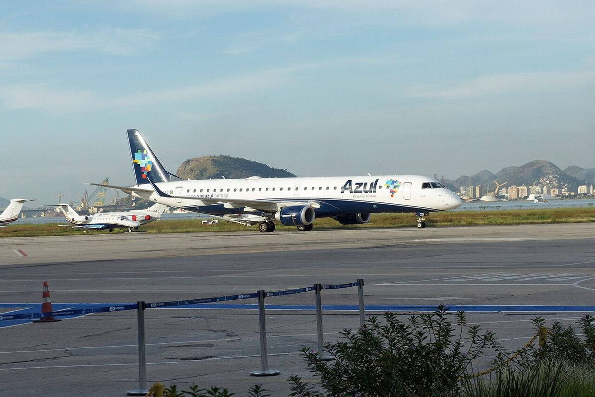 A white Azul Embraer 195 regional jet with a dark blue tail taxiing, with mountains and parked business jets behind.
