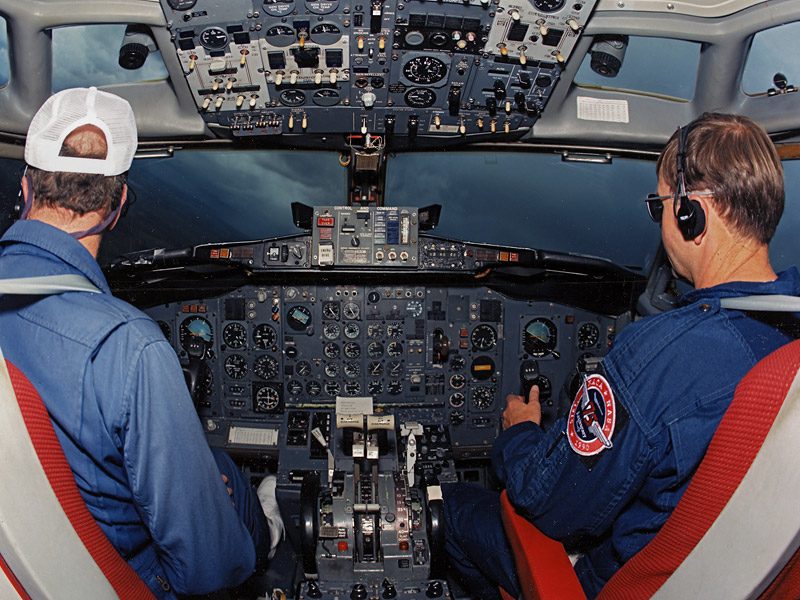 Two pilots seen from behind, sitting side by side in an airliner cockpit and facing a panel of dials, the one on the left wearing a cap.