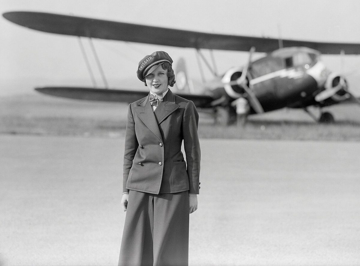 A young woman in a Swissair uniform jacket, trousers and cap standing on an airfield, with a twin-engined biplane behind her.