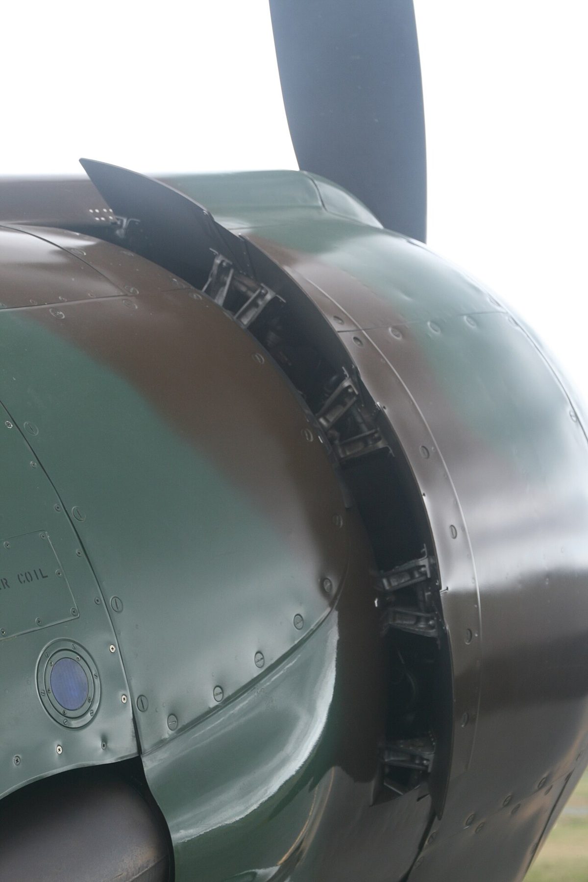 Rear of a round engine cowling on a green and brown painted aircraft, the hinged flaps around its edge standing open.