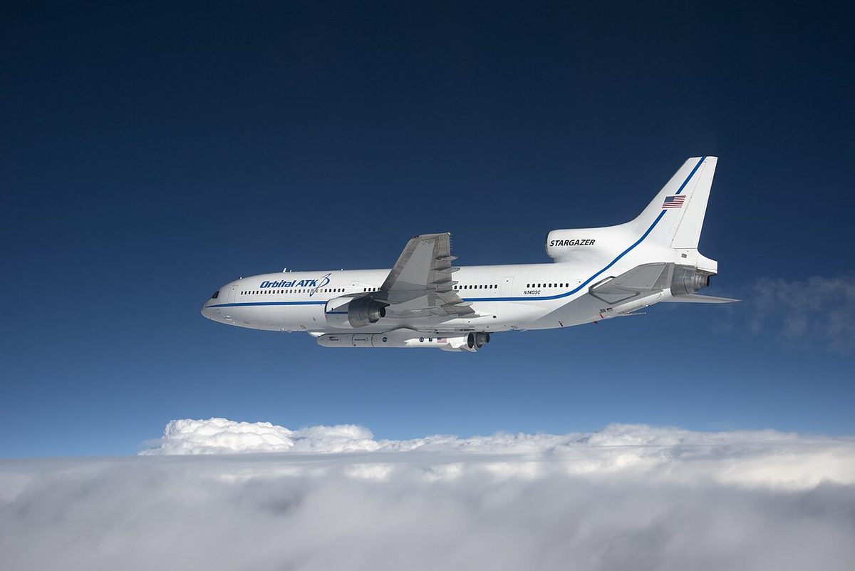 A white three-engined airliner, carrying a winged rocket under its fuselage, flying above an unbroken layer of cloud under a deep blue sky.