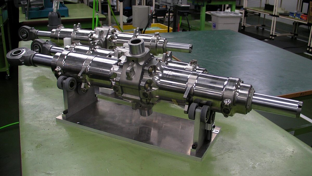 Three polished actuators on a workshop bench, long cylinders with an eye fitting at one end and a central block, the front one held on a stand.