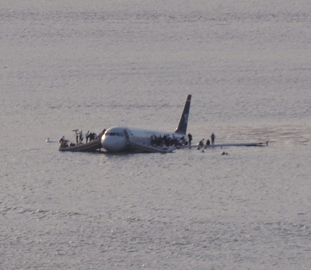 A white airliner floating low in grey river water, a crowd of people standing on both wings and near the forward door.