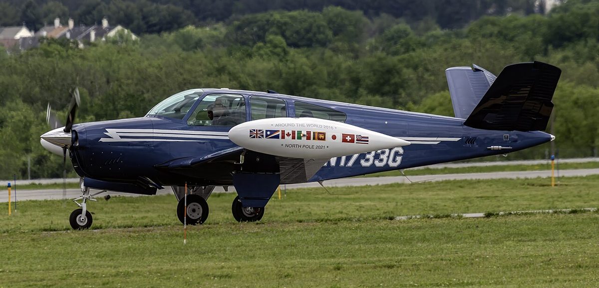 A dark blue Beechcraft Bonanza taxiing past grass, its tail made of two angled surfaces forming a V in place of a separate fin and tailplane.