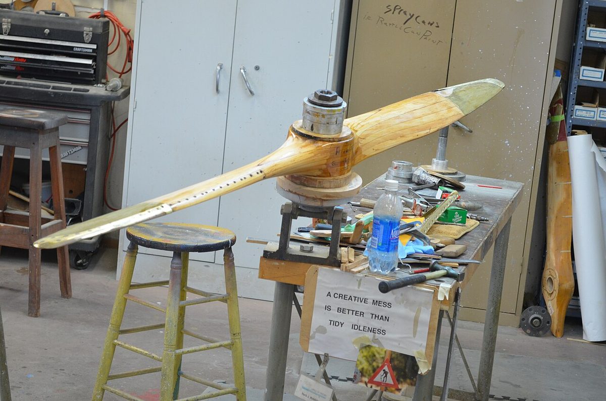 A varnished two-blade wooden propeller mounted on a stand in a workshop, seen at an angle along its length.