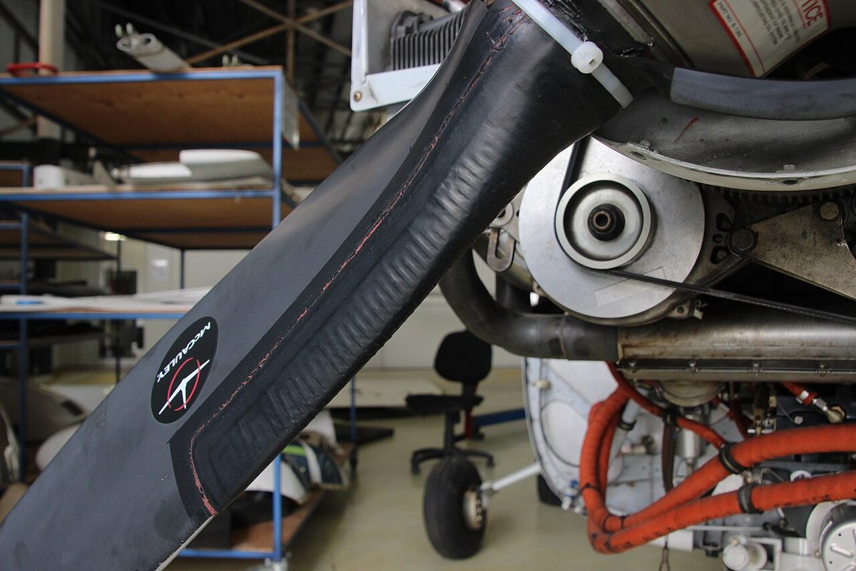 Close-up of a black propeller blade root with a ribbed rubber de-icing boot along its leading edge, engine parts behind it in a workshop.