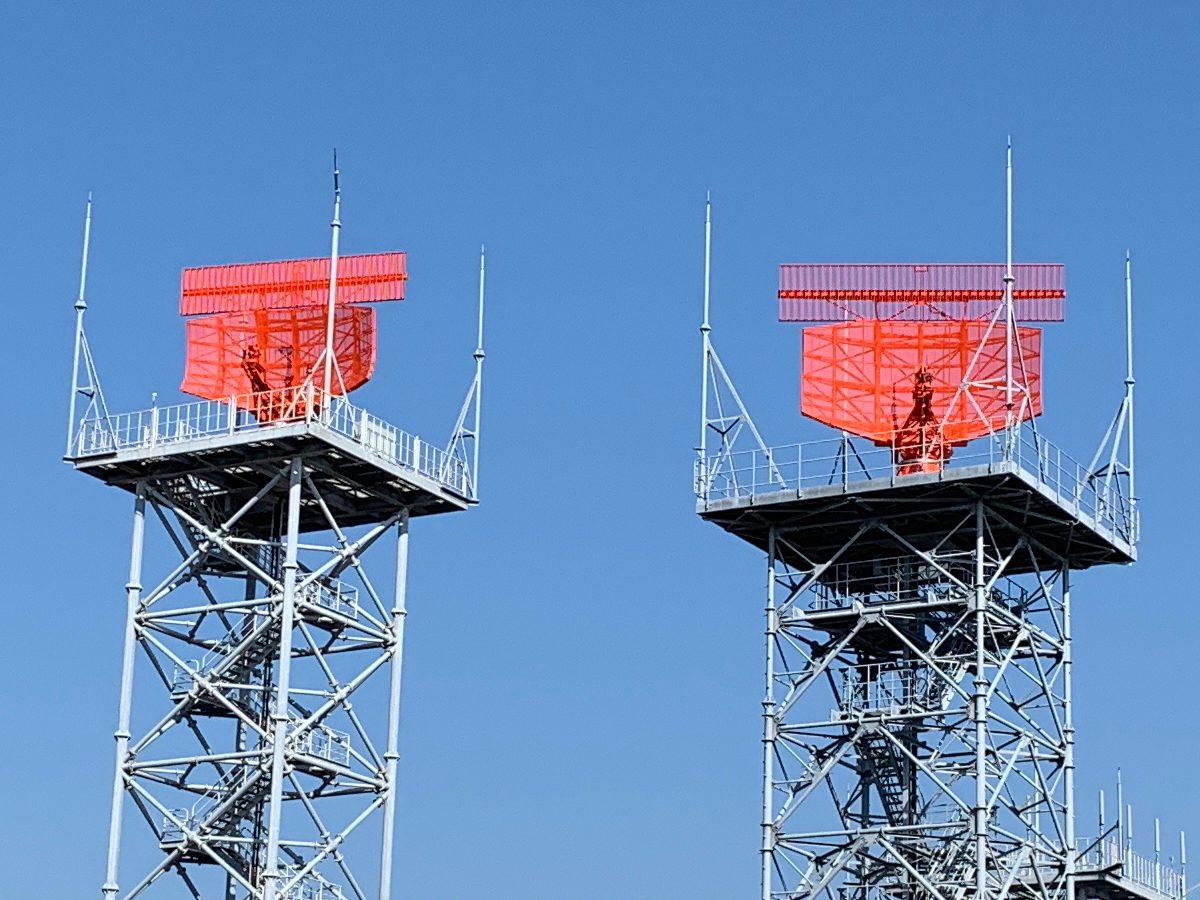 Two lattice towers, each topped by a red curved radar reflector with a flat antenna bar mounted above it, against a blue sky.