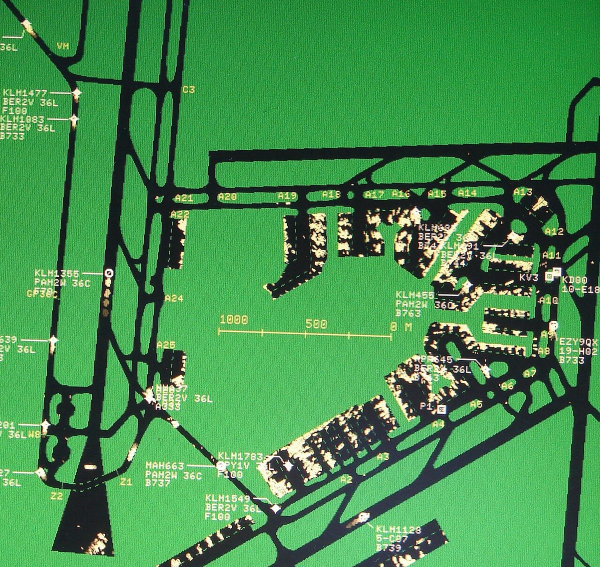 A green surface movement radar screen at Amsterdam Schiphol: taxiways and aprons in black, radar returns from buildings and aircraft in pale yellow, labels with call signs, runways and aircraft types, and a 1000 m scale bar.